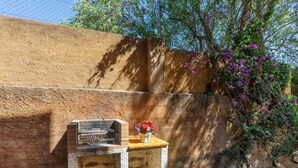 Ein Grillbereich im Freien mit einem Backstein-Grill, einem kleinen Holztisch mit Blumen in einer Vase und einer Betonbank.