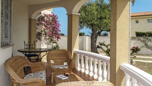 A covered outdoor seating area with wicker furniture, a table, and chairs, overlooking a garden with a tree and flowers.