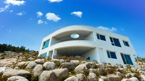 A modern two-story building with a curved balcony, large windows, and a rocky landscape in the foreground. - Sosiega Hue Pension (Goheung)