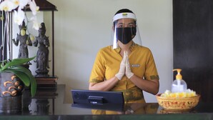 A hotel reception area with a receptionist wearing a yellow uniform, a name tag, and a face shield. There are decorative statues, a vase with flowers, and a bowl of fruit on the counter. - 2 BR Villa in Seminyak, 25sqm Private Pool, 5 mnts walk to shopping area for 6 (Seminyak)
