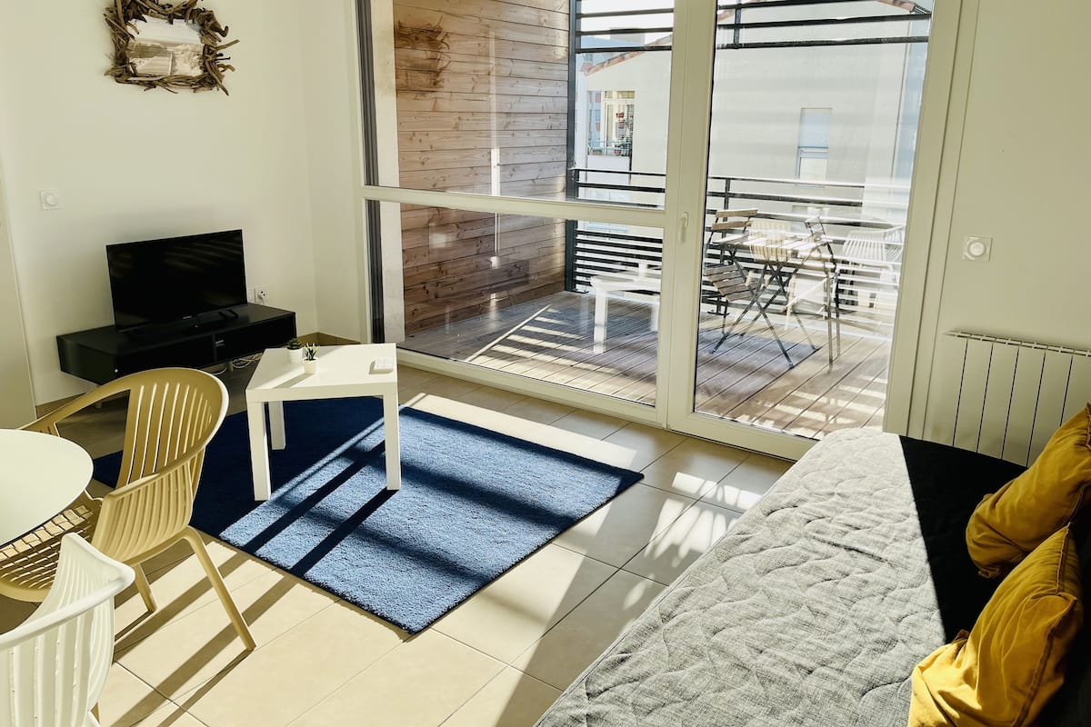 A modern living room with a sofa, a TV, a dining table with chairs, and a balcony with a view of the outside.