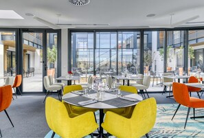 A modern dining area with round tables, yellow chairs, and large windows offering a view of the outside. - Les Patios de Saint-Pierre (Palavas-les-Flots)