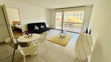 Ein modernes Wohnzimmer mit einer schwarzen Couch, weißen Sesseln, einem kleinen runden Tisch und einem großen Fenster mit Blick auf einen Balkon.