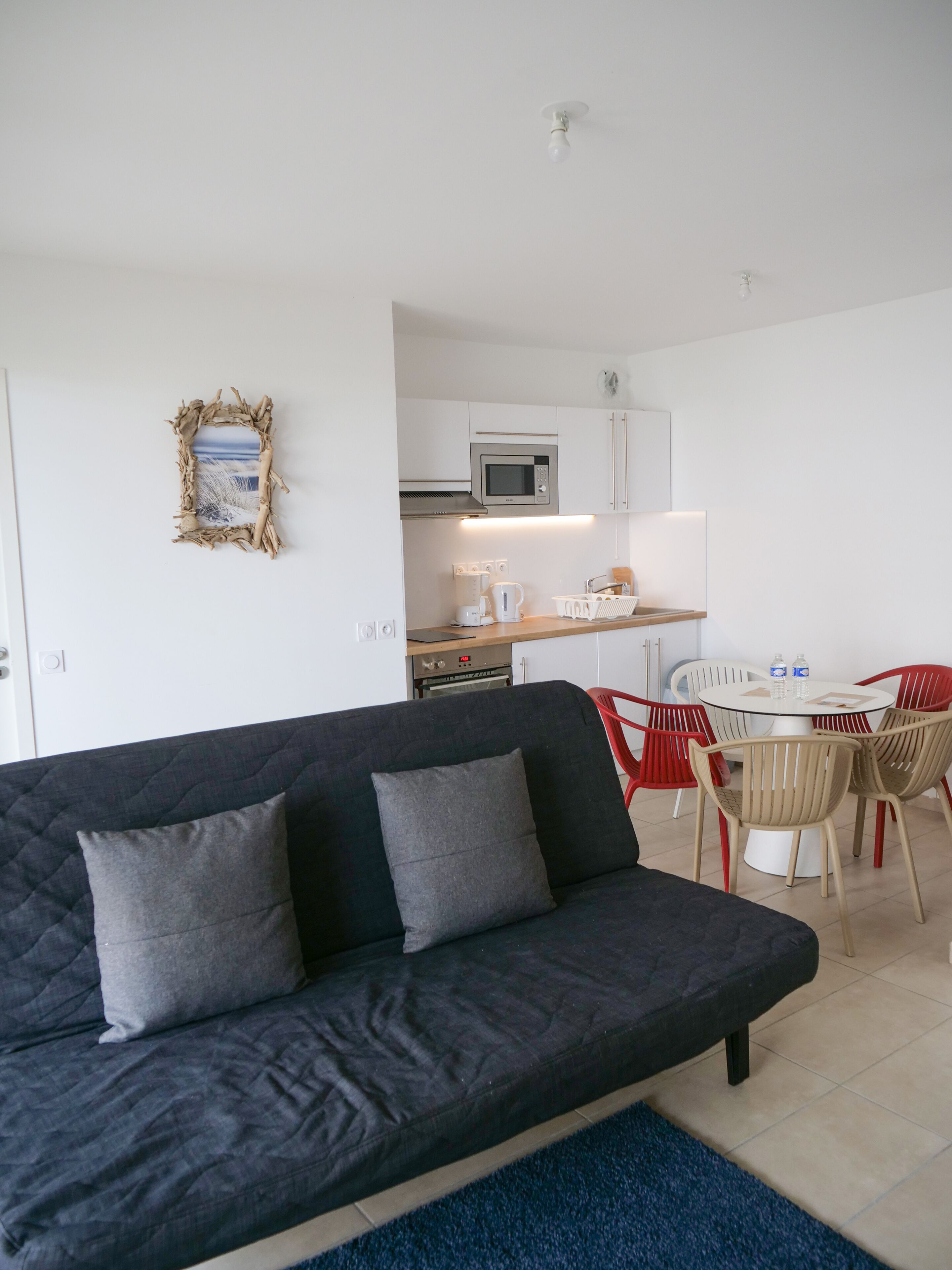 A modern living room with a dark sofa, a dining area with red and white chairs, a white kitchen with a microwave, and a framed picture on the wall.