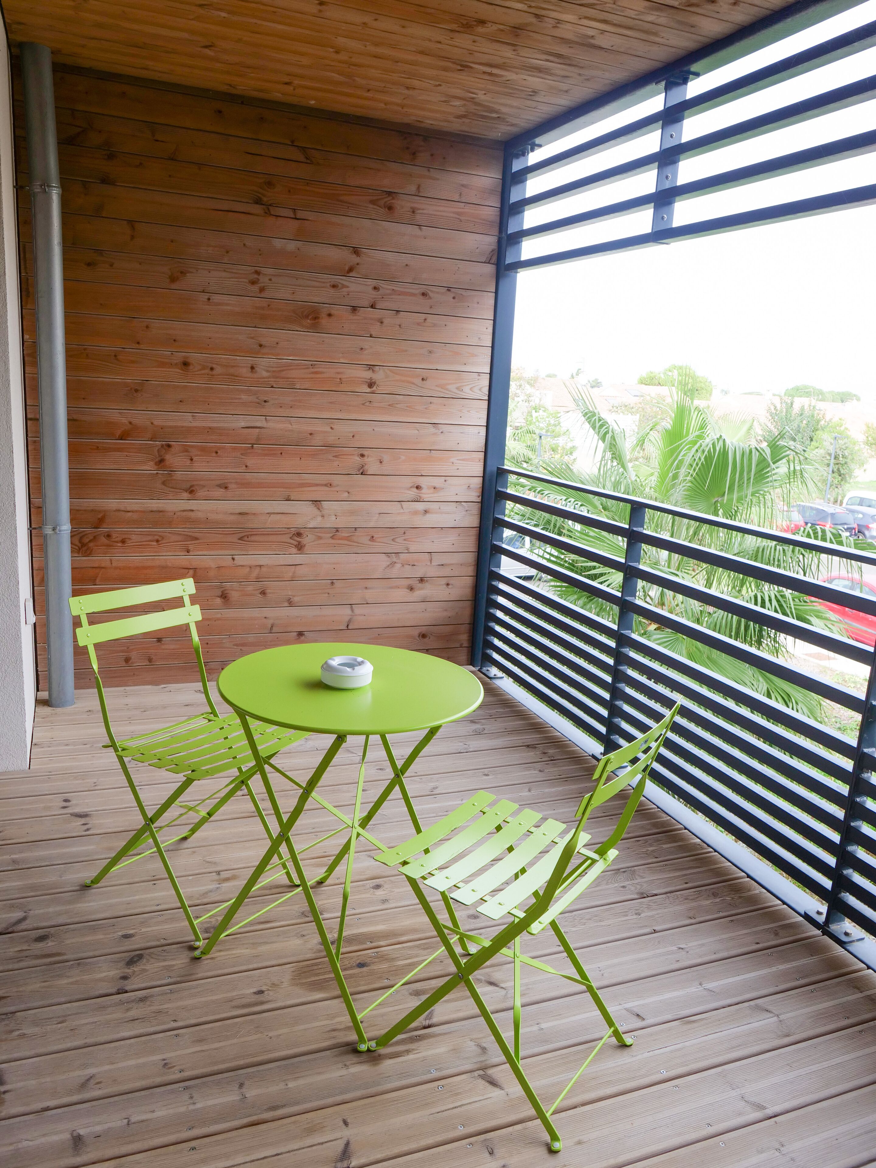 A wooden deck with a round table and two chairs, a metal railing, and a view of greenery and parked cars.