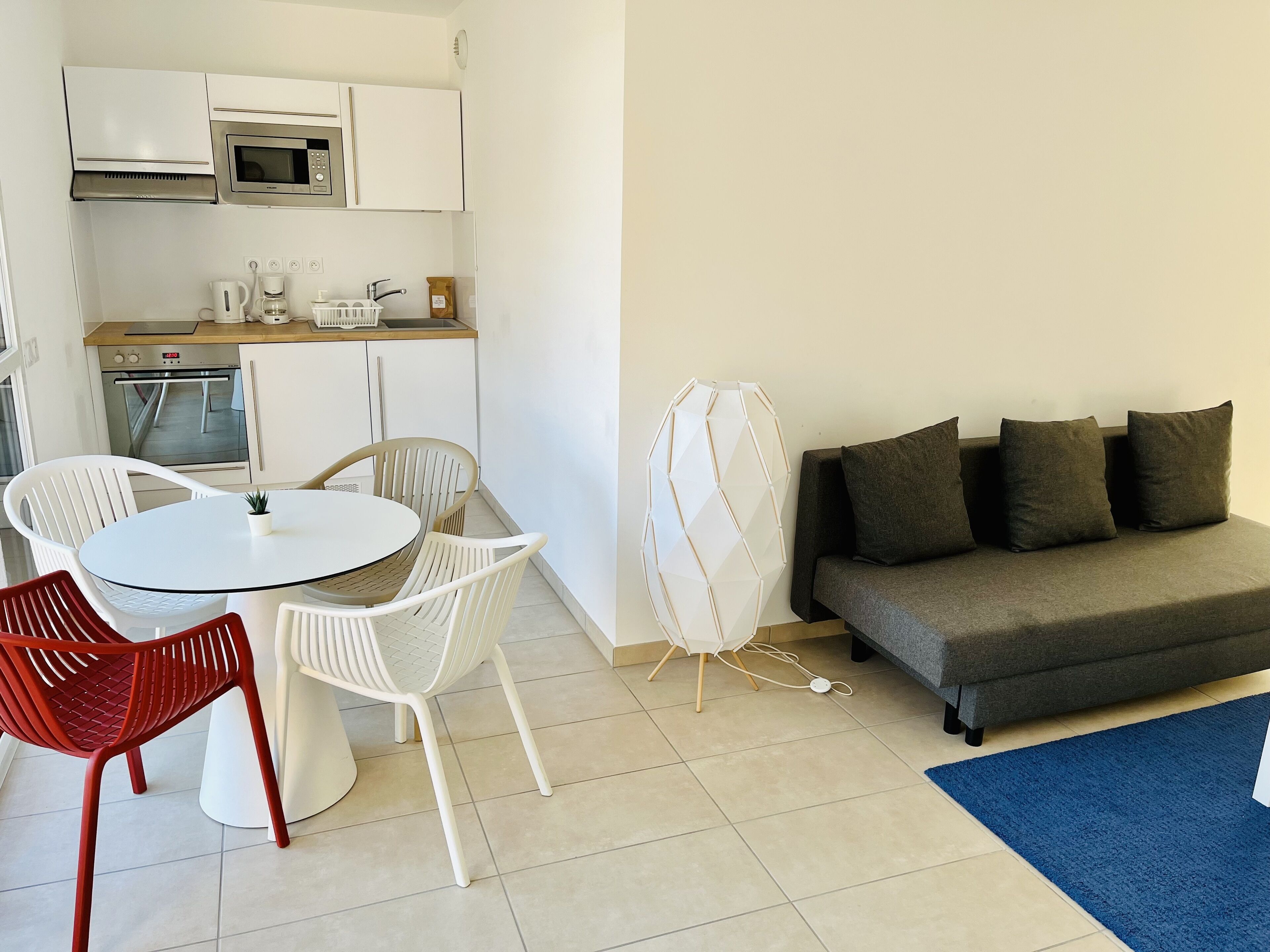 Una cocina moderna con armarios blancos, microondas y cafetera. Hay una mesa de comedor con sillas, un sofá y una alfombra azul.
