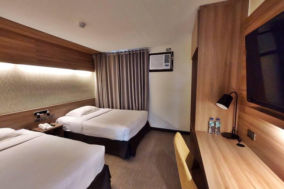A hotel room with two beds, a desk, a TV, a lamp, and a window with curtains.