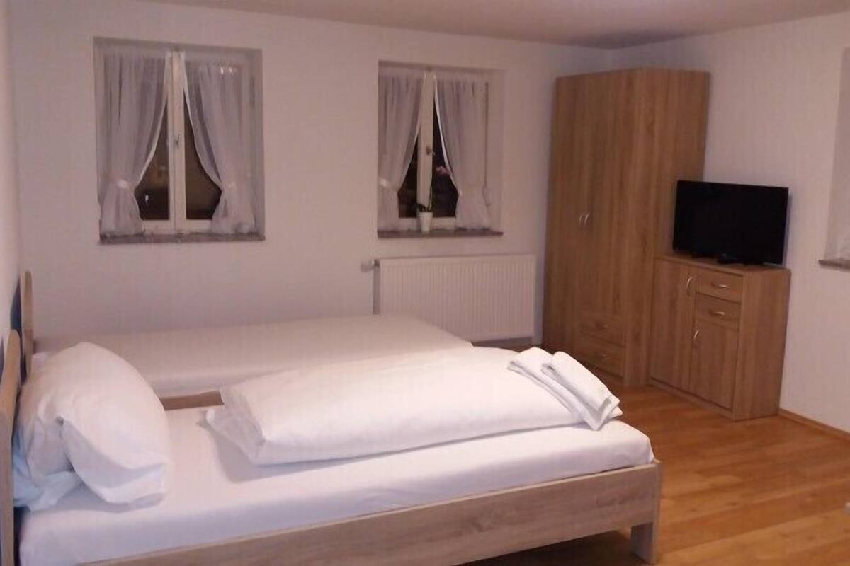 A bedroom with a wooden bed, a TV on a wooden cabinet, and two windows with curtains.