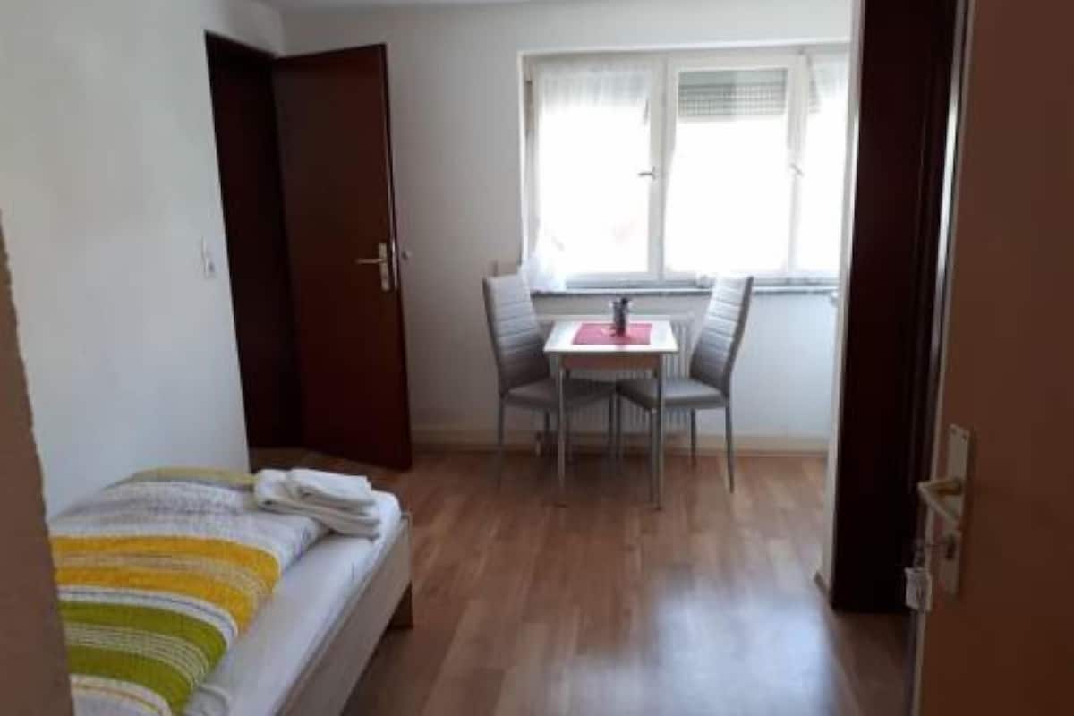 A small, tidy room with a bed, a table with two chairs, and a window with blinds.