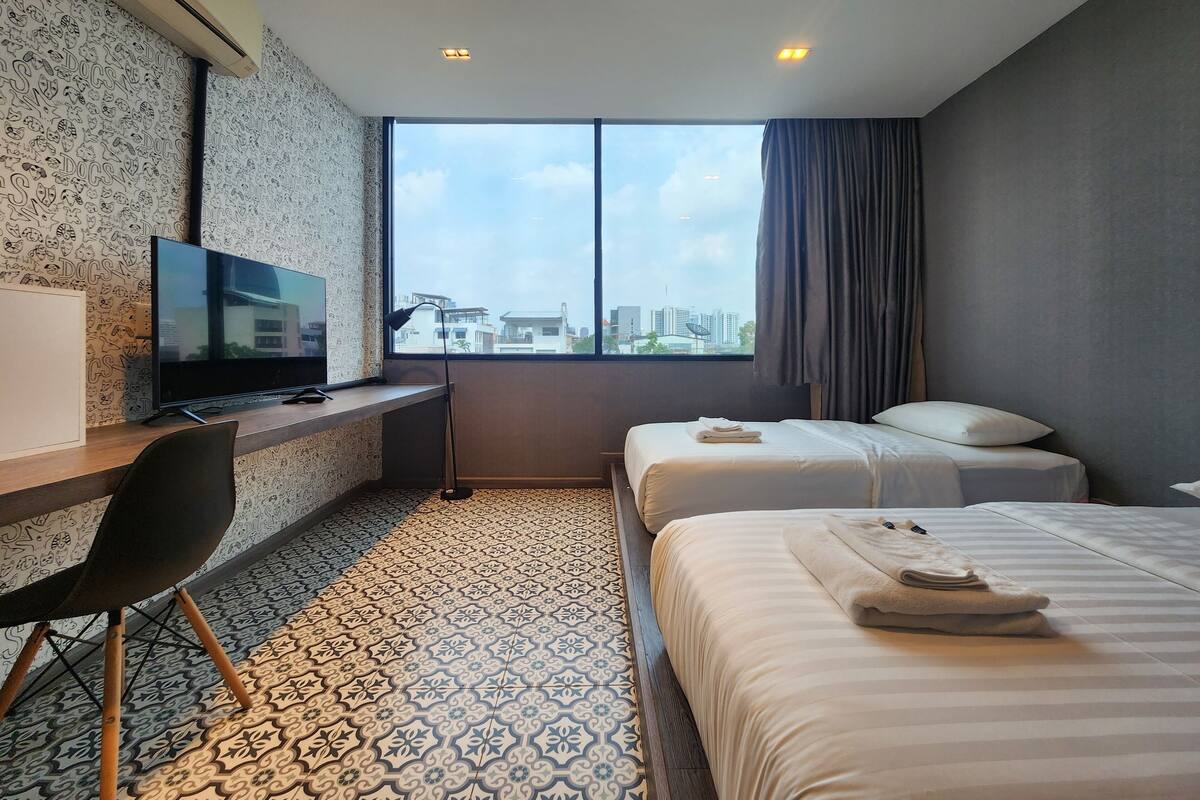 Quarto de hotel com uma grande janela, uma mesa com televisão, duas camas e piso de azulejos estampados.