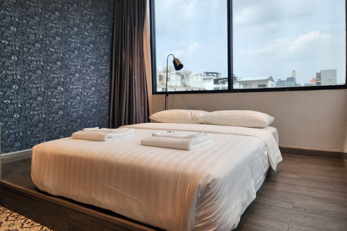Quarto de hotel com cama grande, janela com vista para a cidade, parede escura com padrão e luminária moderna.
