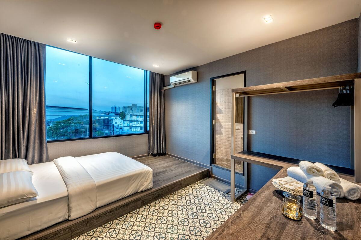 Quarto de hotel moderno com uma janela grande, uma cama, uma mesinha de apoio com artigos de higiene pessoal e um espelho.