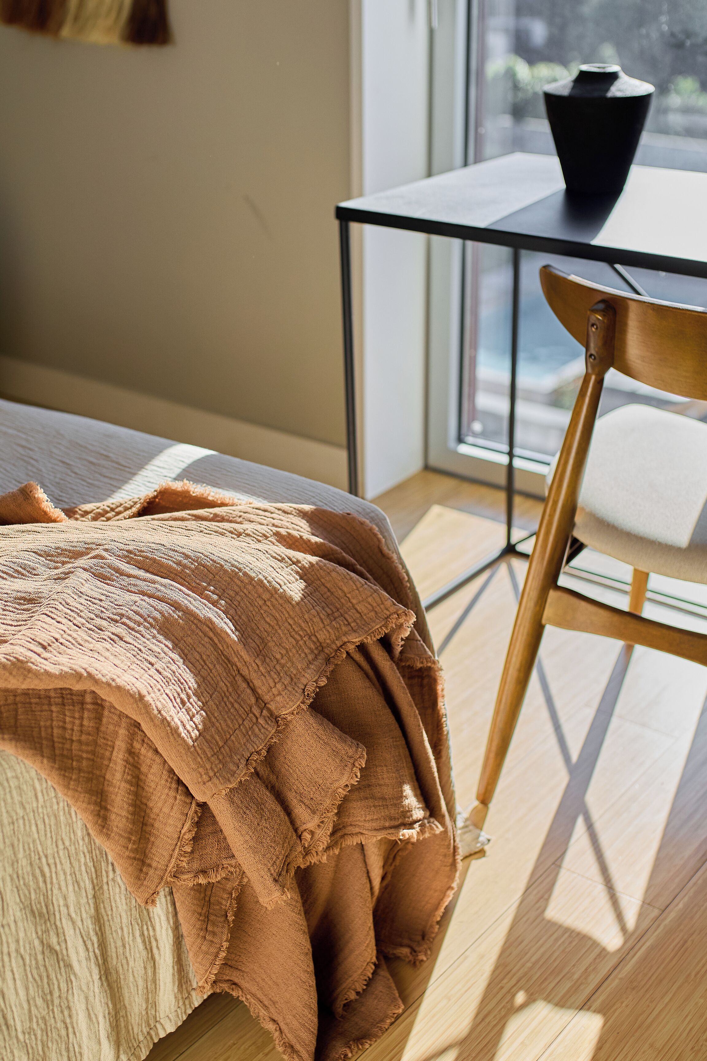 Une pièce avec un lit recouvert d’une couverture à motifs, une chaise en bois, une table sur laquelle se trouve un vase noir, et une fenêtre laissant entrer la lumière naturelle.