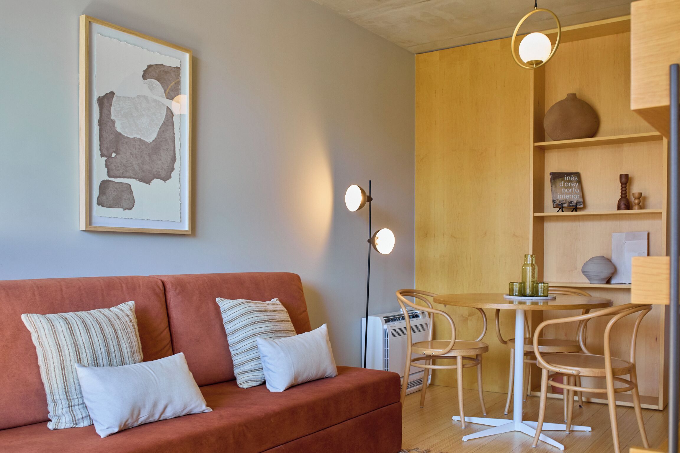 Un salon moderne avec un canapé rouge, des coussins blancs et à rayures, une table ronde avec des chaises et un luminaire fixé au mur.