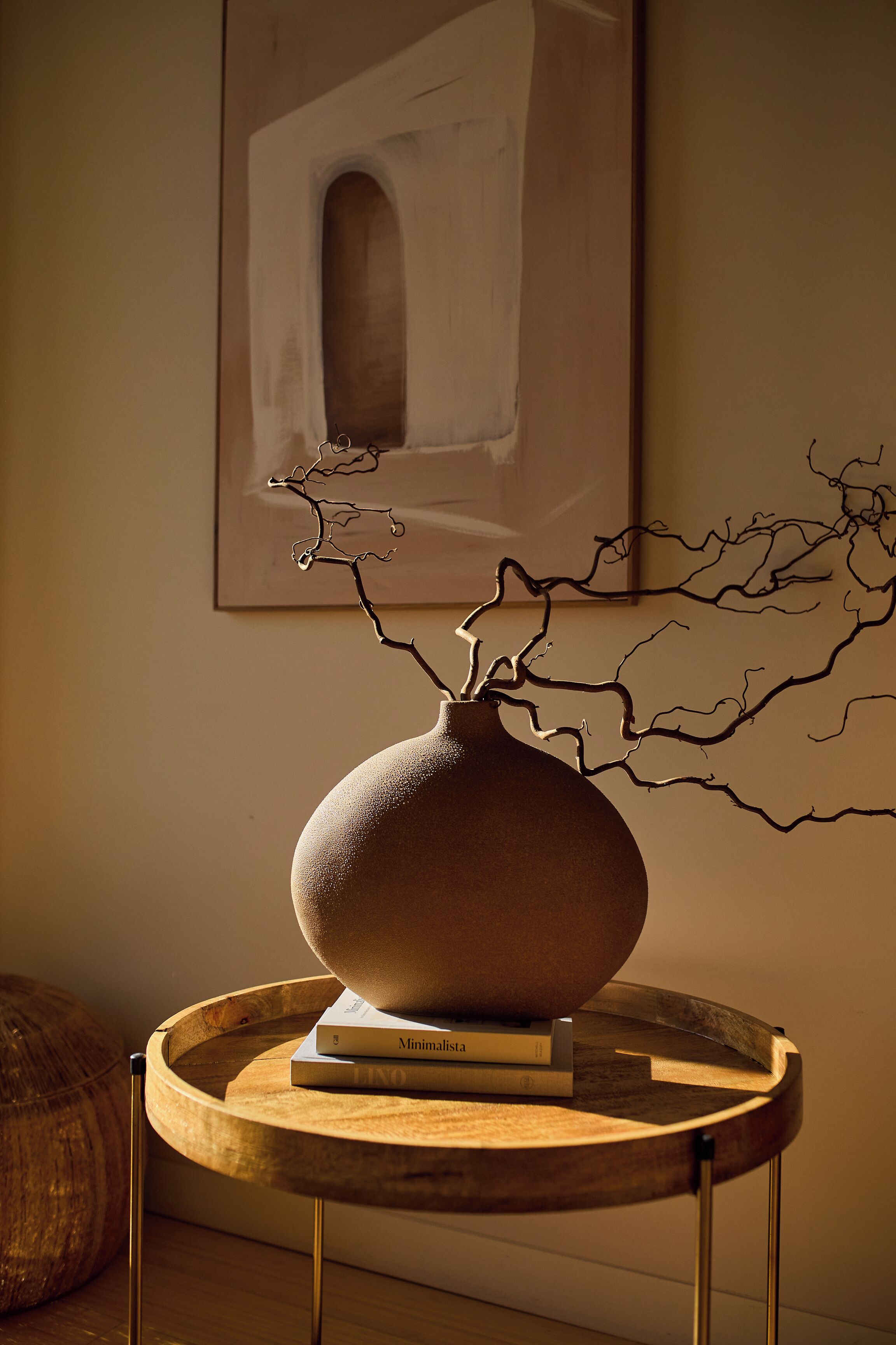 Een ronde houten tafel met een keramieken vaas en boeken, een houten kruk, en een kunstwerk in een lijst aan de muur.