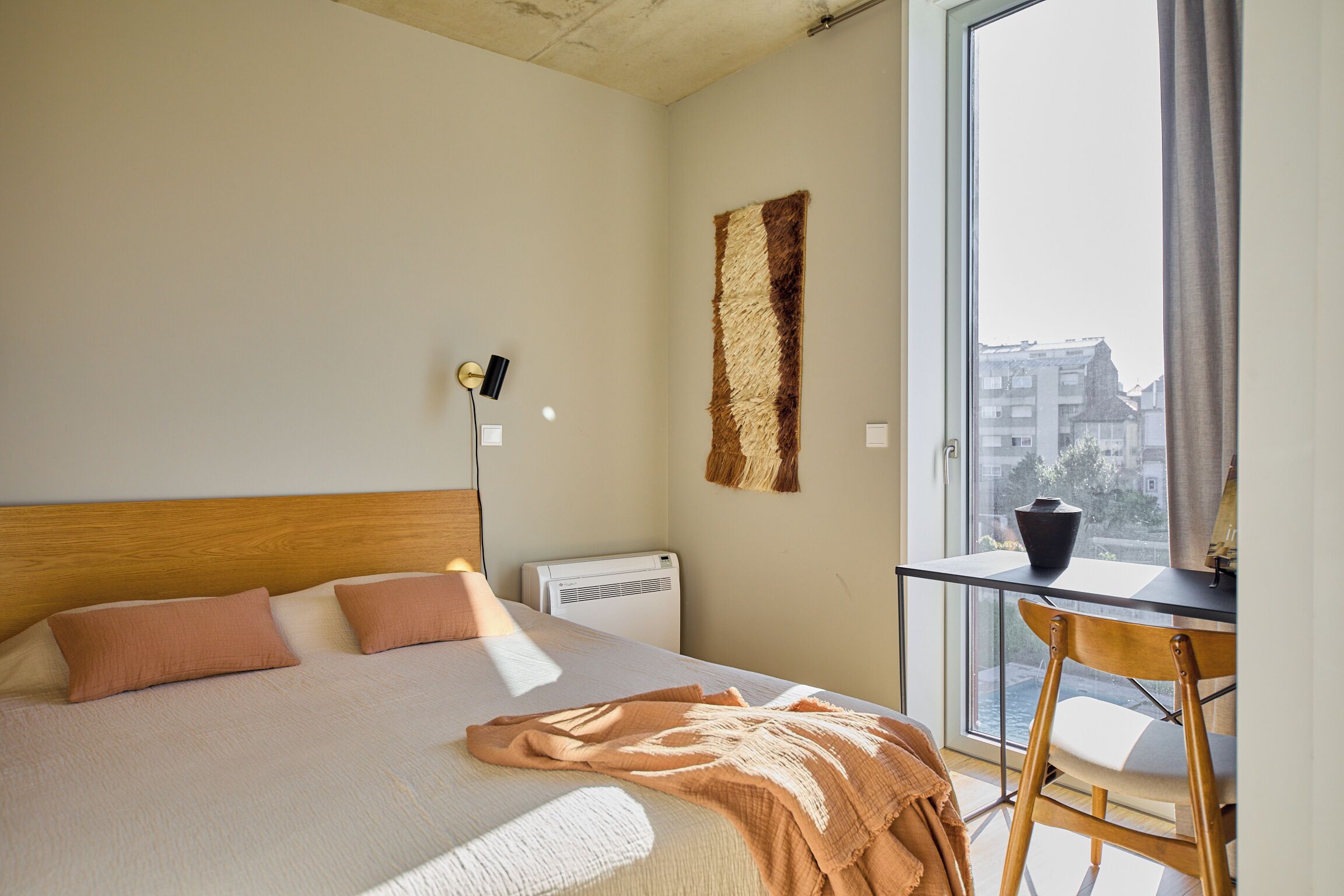 Ein Schlafzimmer mit Bett, Schreibtisch und einem Fenster mit Blick auf Gebäude.