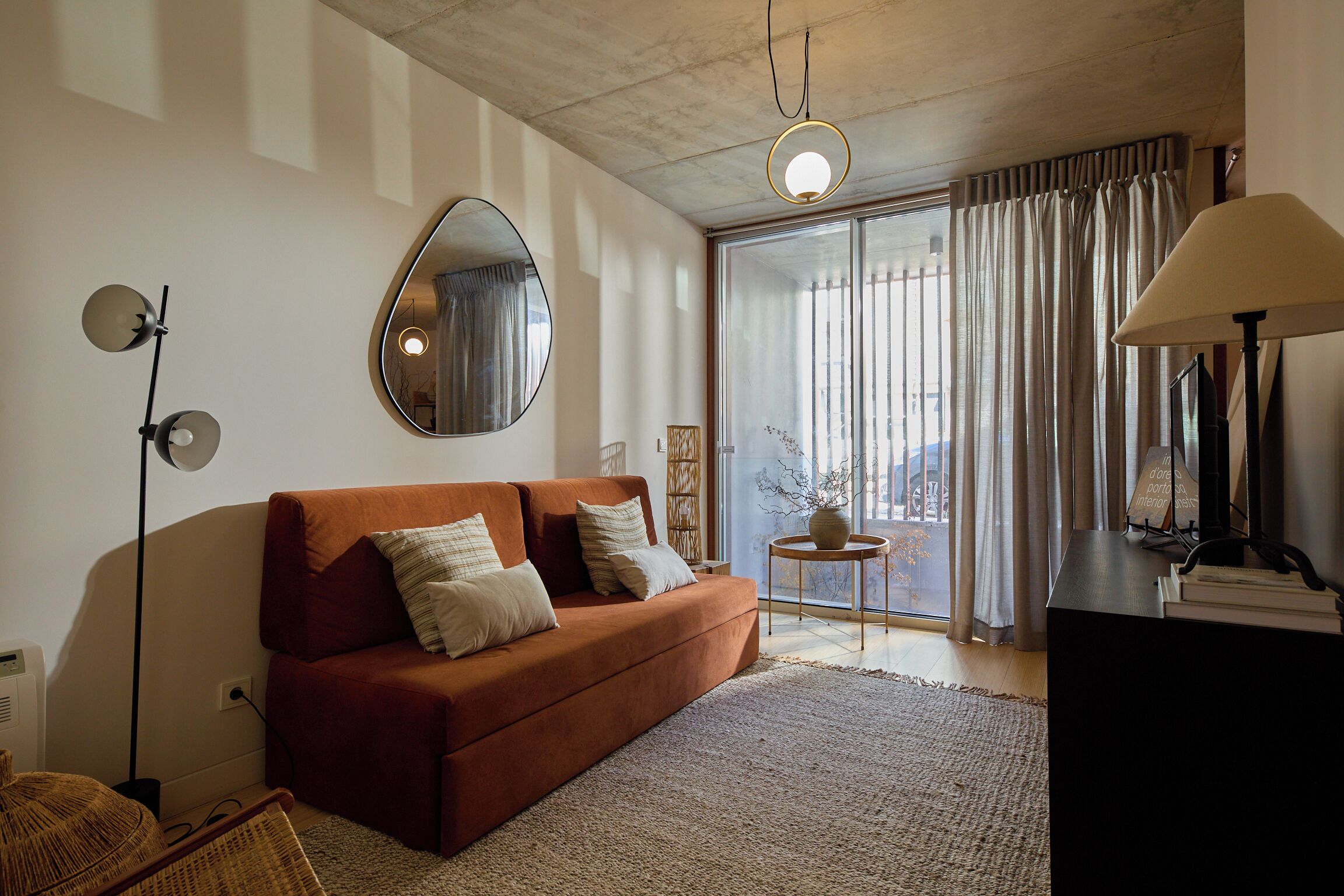 A modern living room with a sofa, a round mirror, a floor lamp, and a view through a large window with curtains.