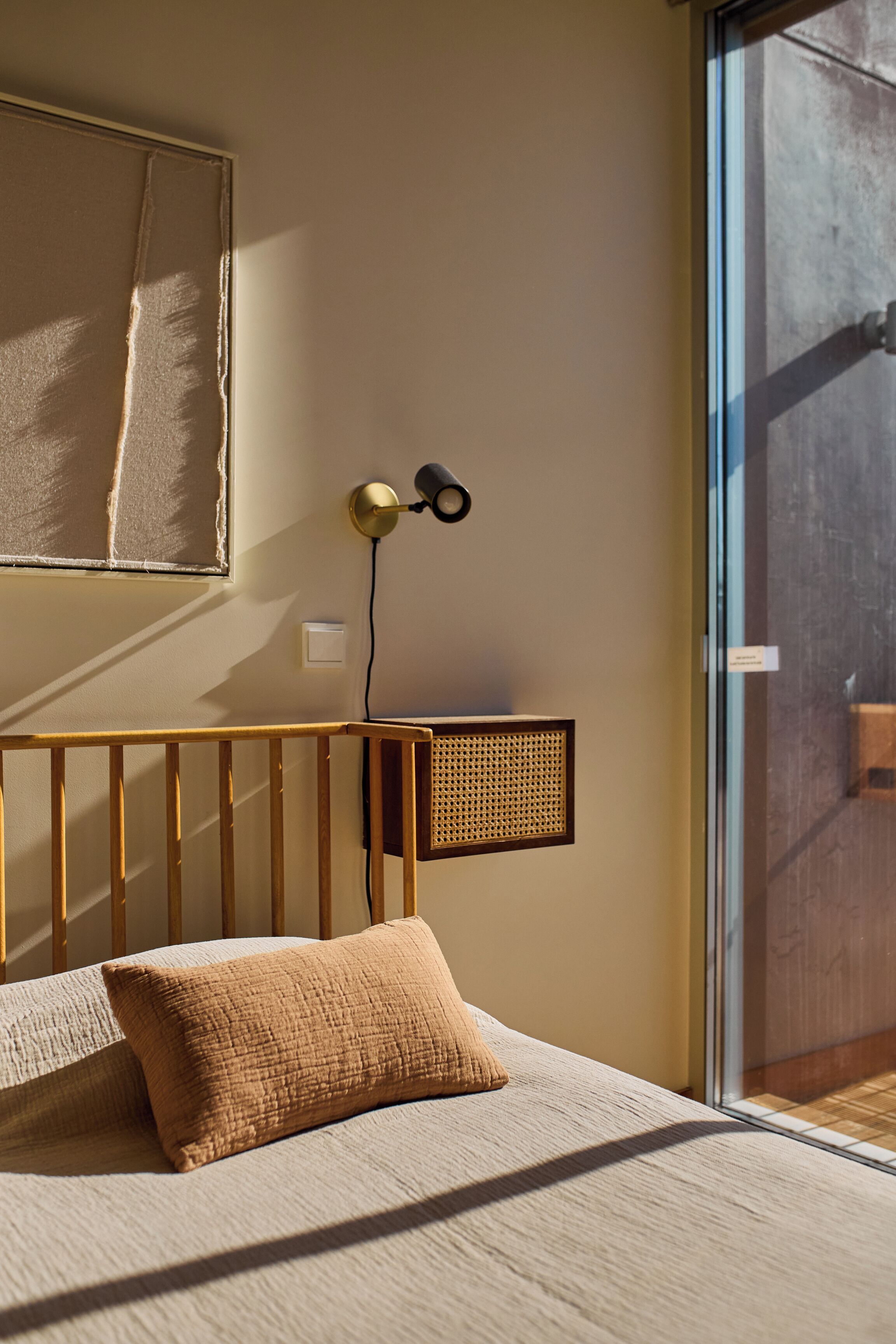 Een slaapkamer met een houten hoofdbord, een bed met beige beddengoed, een wandlamp en een raam met uitzicht.
