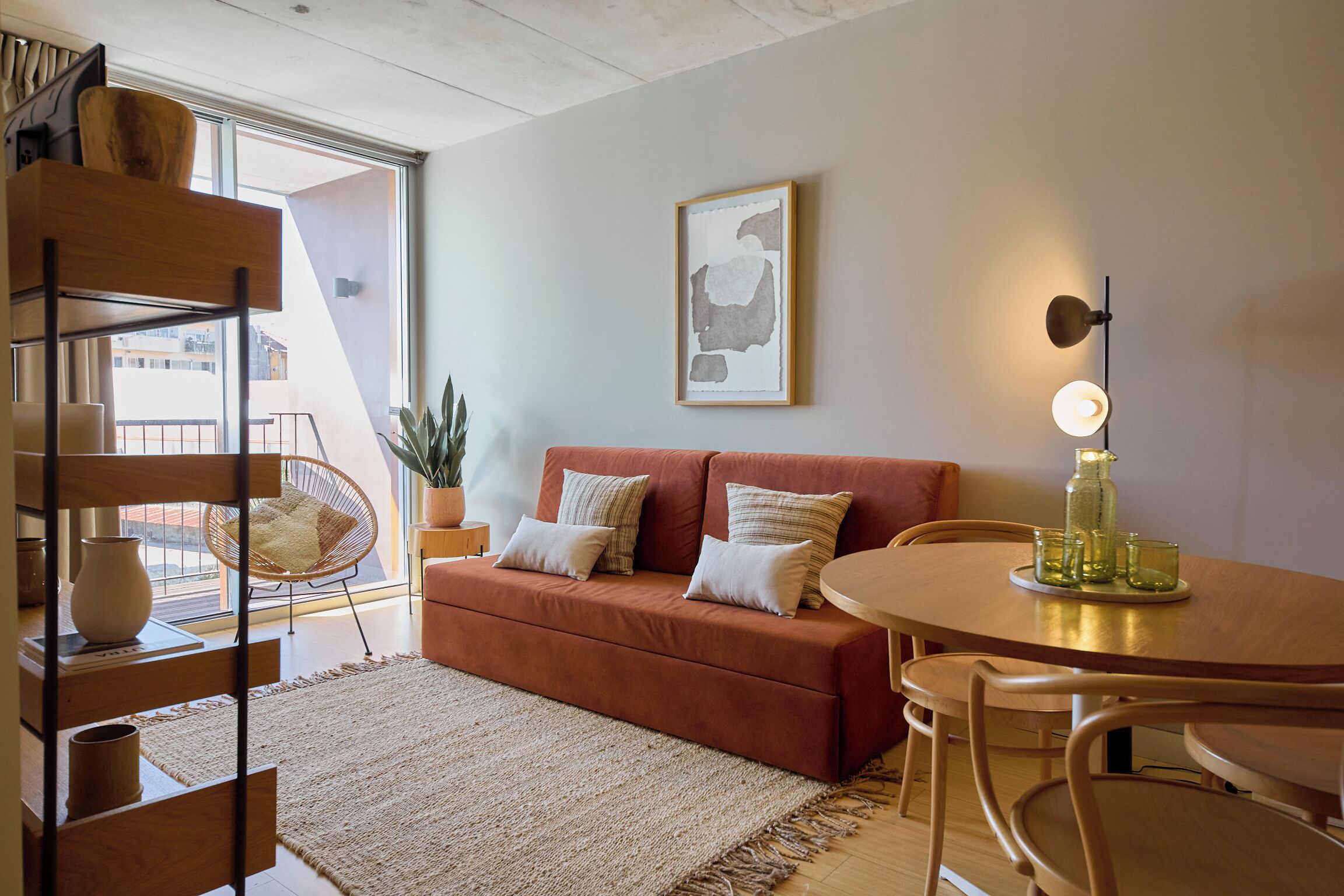 A modern living room with a brown sofa, a round wooden table, and a chair. There is a large window providing natural light and a view of the outside.
