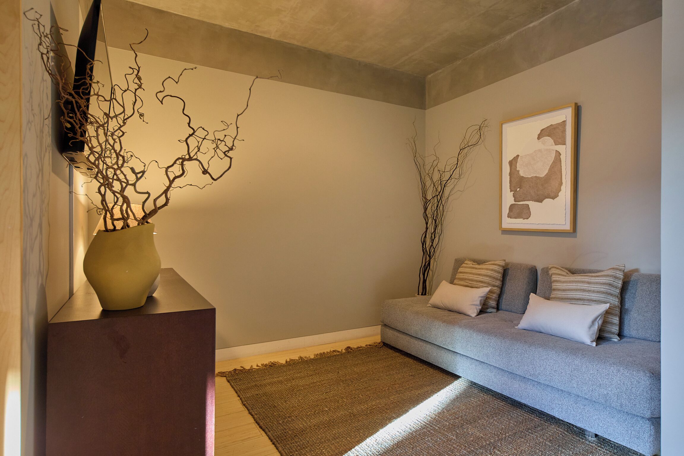 A living room with a grey sofa, a brown rug, and a vase with branches on a wooden stand.