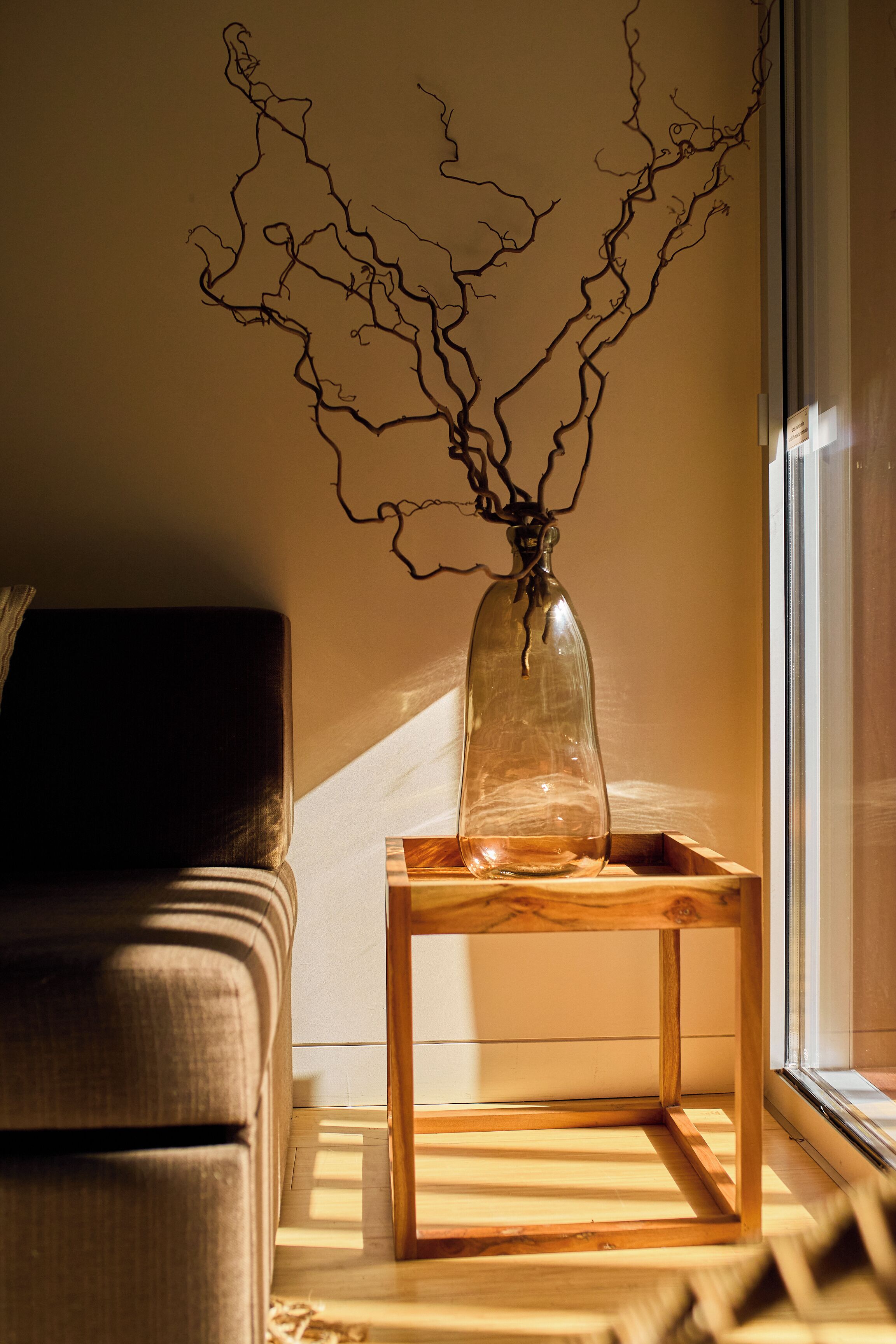 Una mesa de madera con un jarrón de cristal que contiene ramas, ubicada cerca de una ventana donde la luz del sol proyecta sombras sobre el suelo.