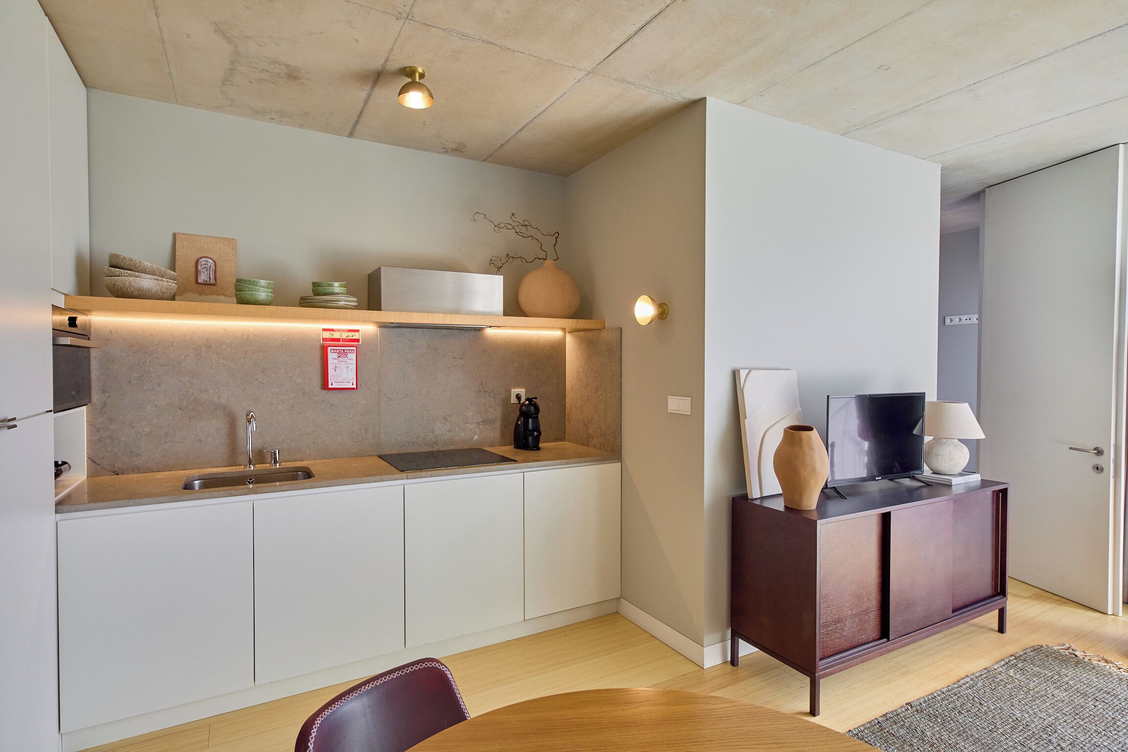 A compact kitchen with a sink, countertop, and cabinets. There is a dining table with a chair. A cabinet with decorative items and a television is visible.