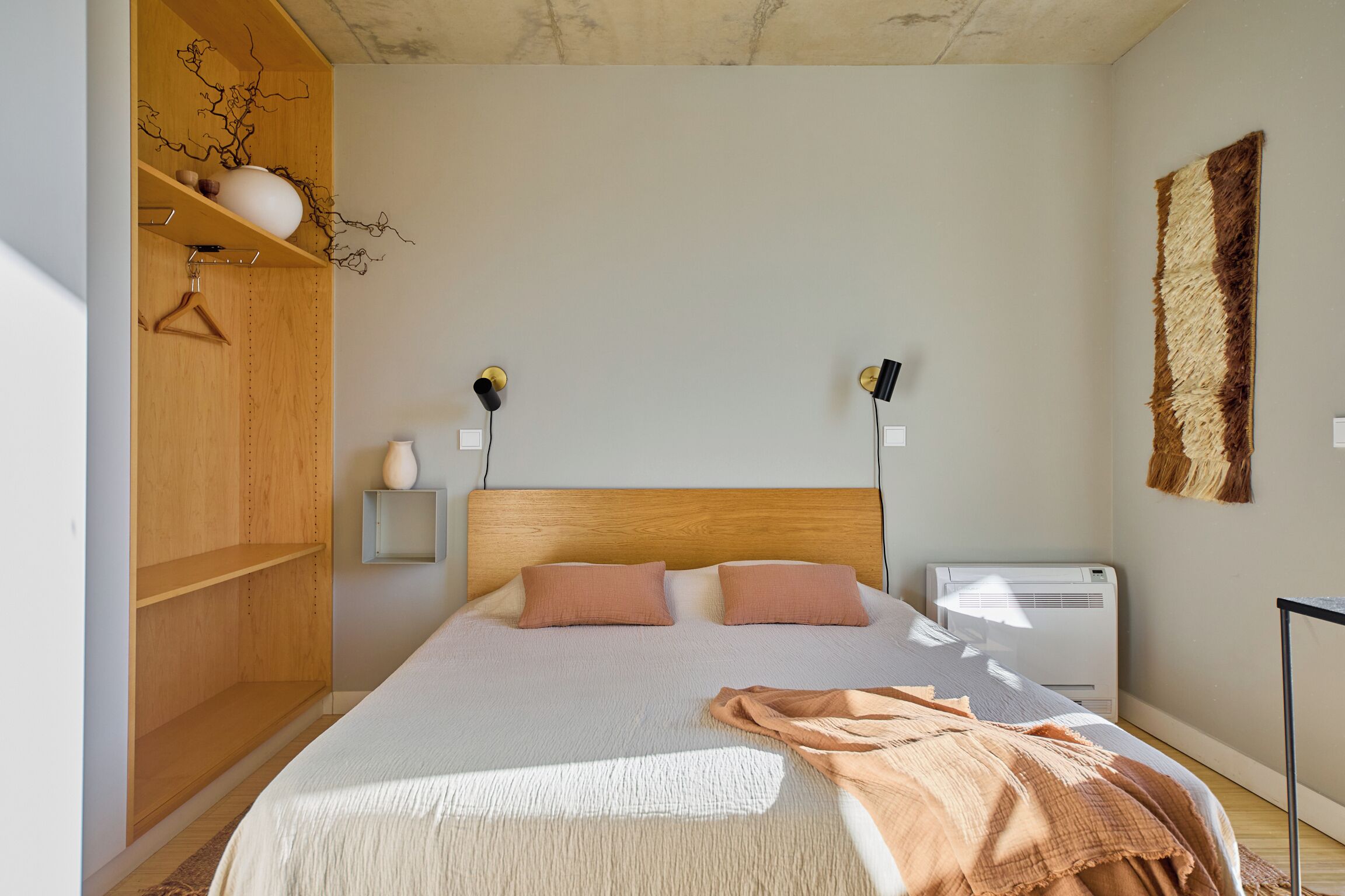 Quarto com cabeceira de madeira, duas mesinhas de cabeceira, cama com edredom branco e bege, e planta pendurada.
