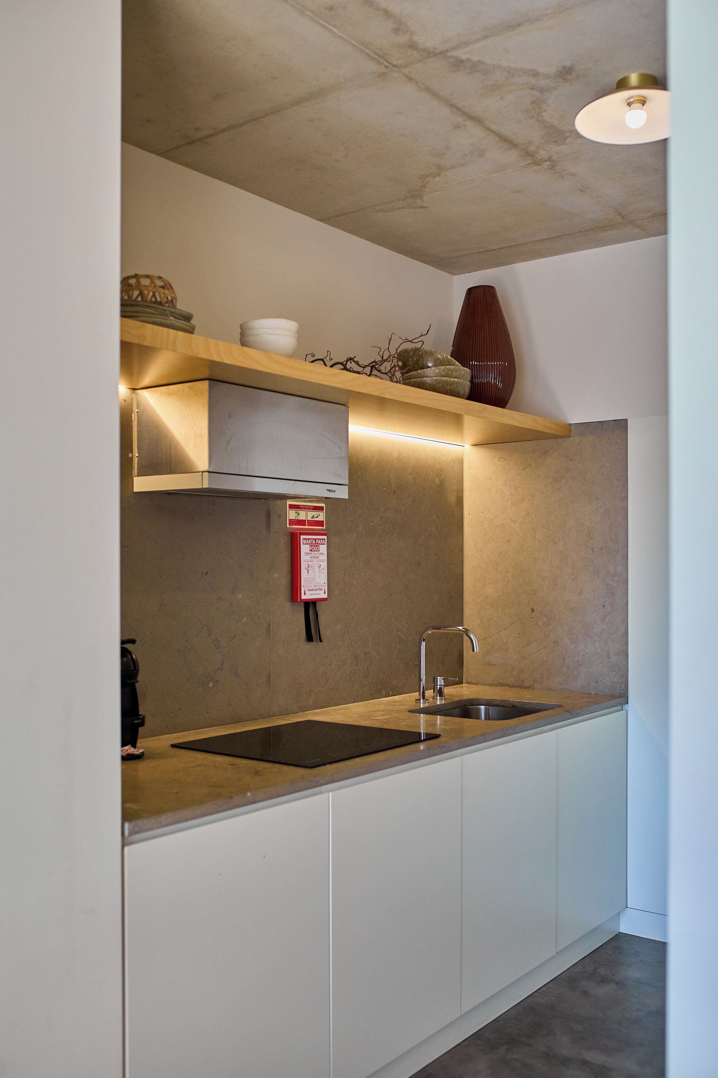 A modern kitchen with a stainless steel sink, white cabinets, a wooden shelf with decorative items, and a fire extinguisher.