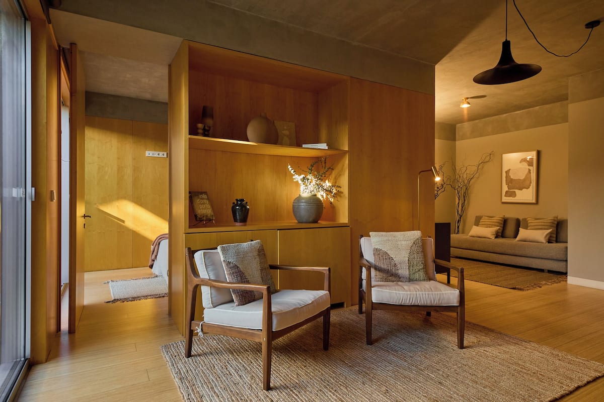 Una sala moderna con piso de madera, dos sillones, un sofá, una estantería y una lámpara colgante.