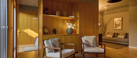 A modern living room with wooden flooring, two armchairs, a sofa, a bookshelf, and a hanging light fixture.