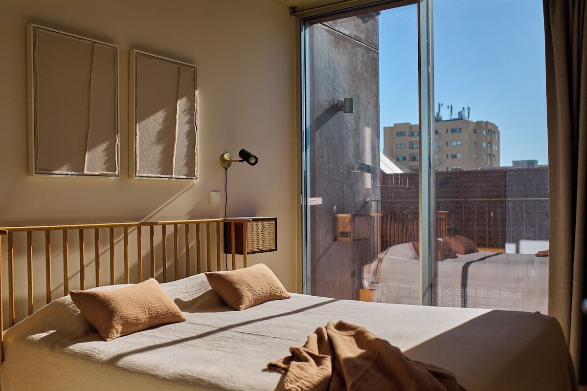 Habitación de hotel con una cama grande, dos almohadas, una mesita de noche, una lámpara y vistas a edificios a través de un ventanal amplio.