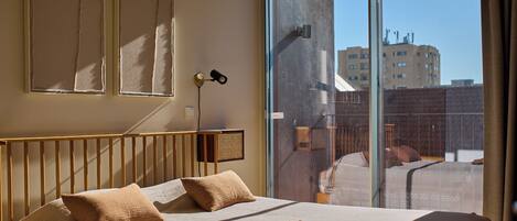 A hotel room with a large bed, two pillows, a bedside table, a lamp, and a view of buildings through a large window.