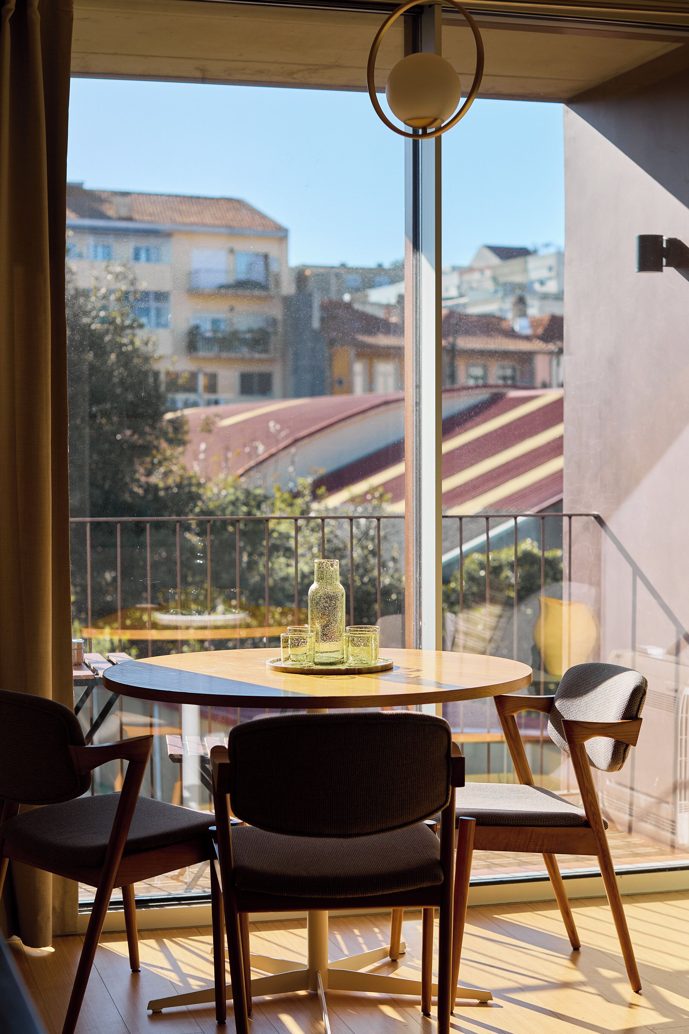 A balcony with a round table and four chairs, overlooking a residential area with houses and a red-roofed building.