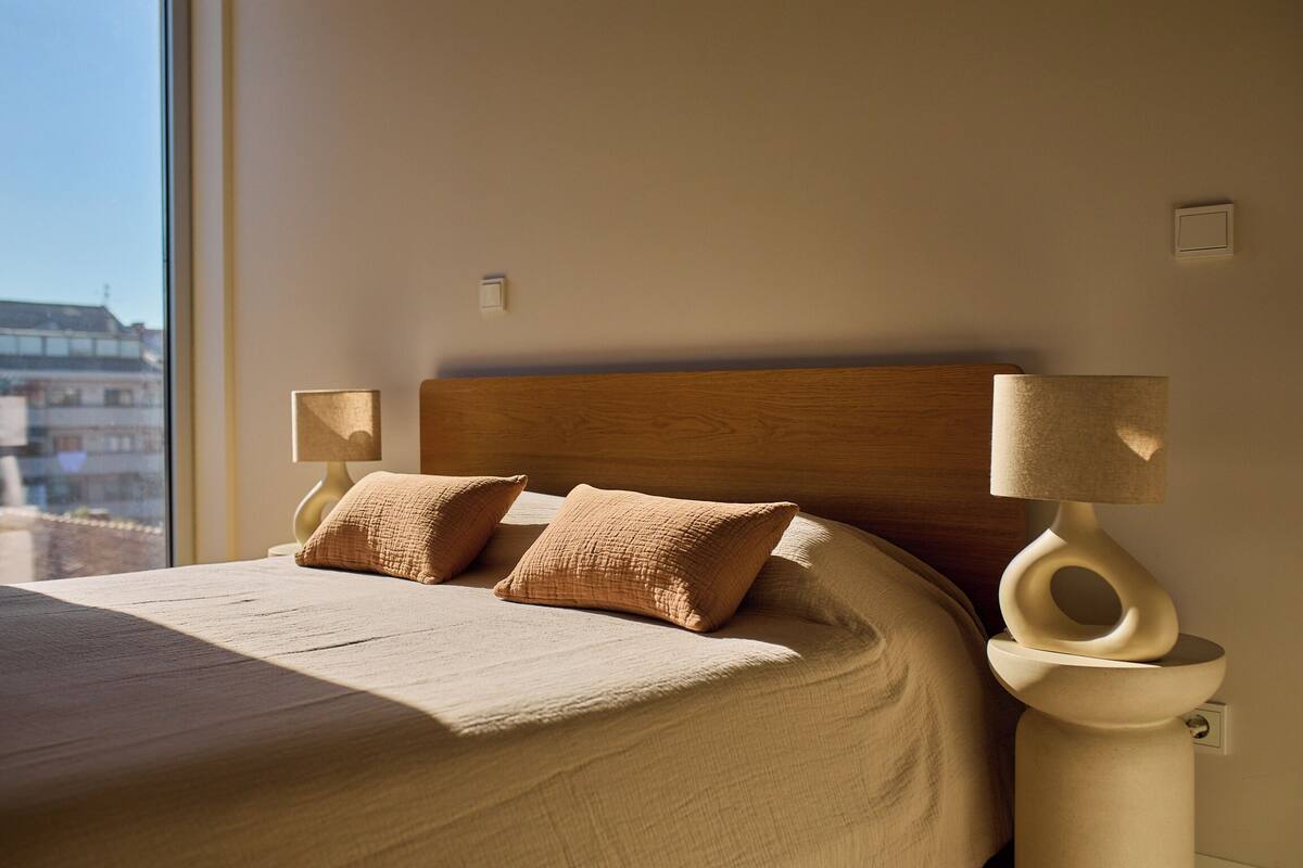 Un dormitorio con una cama grande, dos lámparas de noche y vistas a edificios a través de una ventana.