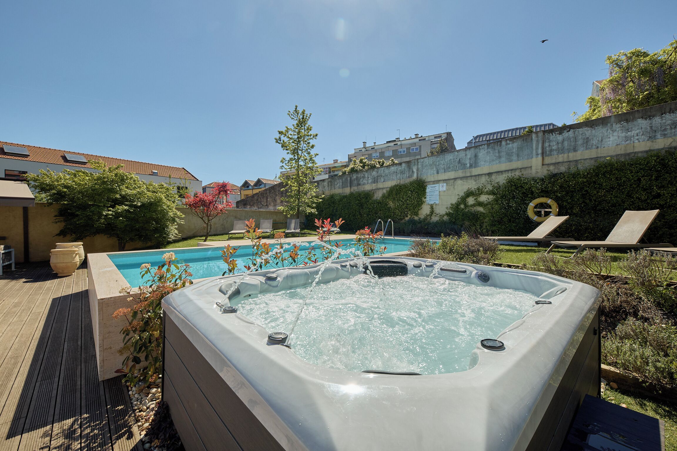 Ett bubbelbad omgivet av ett trädäck, en swimmingpool och en trädgård med växter och en mur.