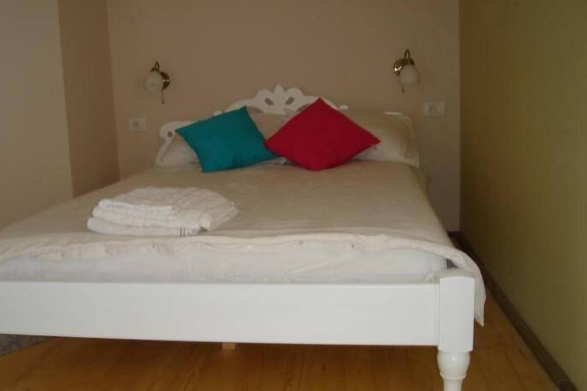 A neatly made bed with white bedding and two pillows, one teal and one red, in a room with wooden flooring and beige walls.
