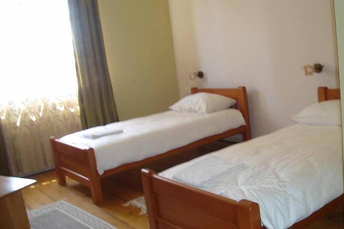Two wooden single beds with white linens in a room with a wooden floor and a window with curtains.
