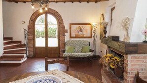 Un salón acogedor con chimenea, un sofá con cojines verdes, una mesa de comedor con un plato decorativo y una escalera que lleva a un espacio al aire libre.