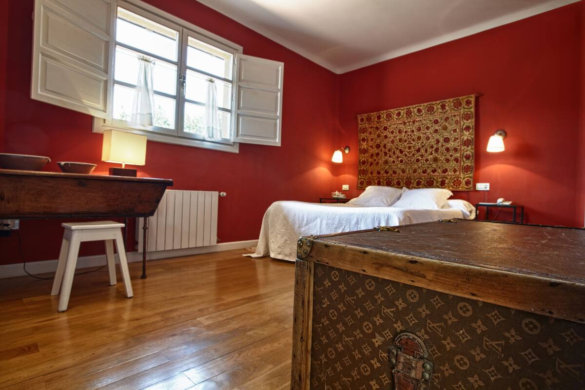 A room with a wooden floor, a bed with white bedding, a red wall, a tapestry, and a lamp.