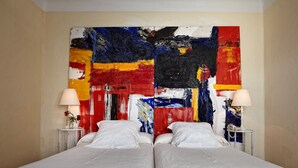 Habitación de hotel con un cuadro abstracto grande en la pared, dos camas, mesitas de noche con lámparas y un jarrón con flores.