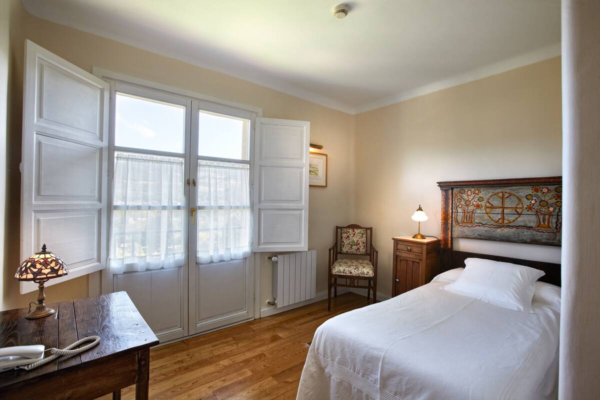 A bedroom with a bed, a chair, a desk with a lamp, and a window with curtains.