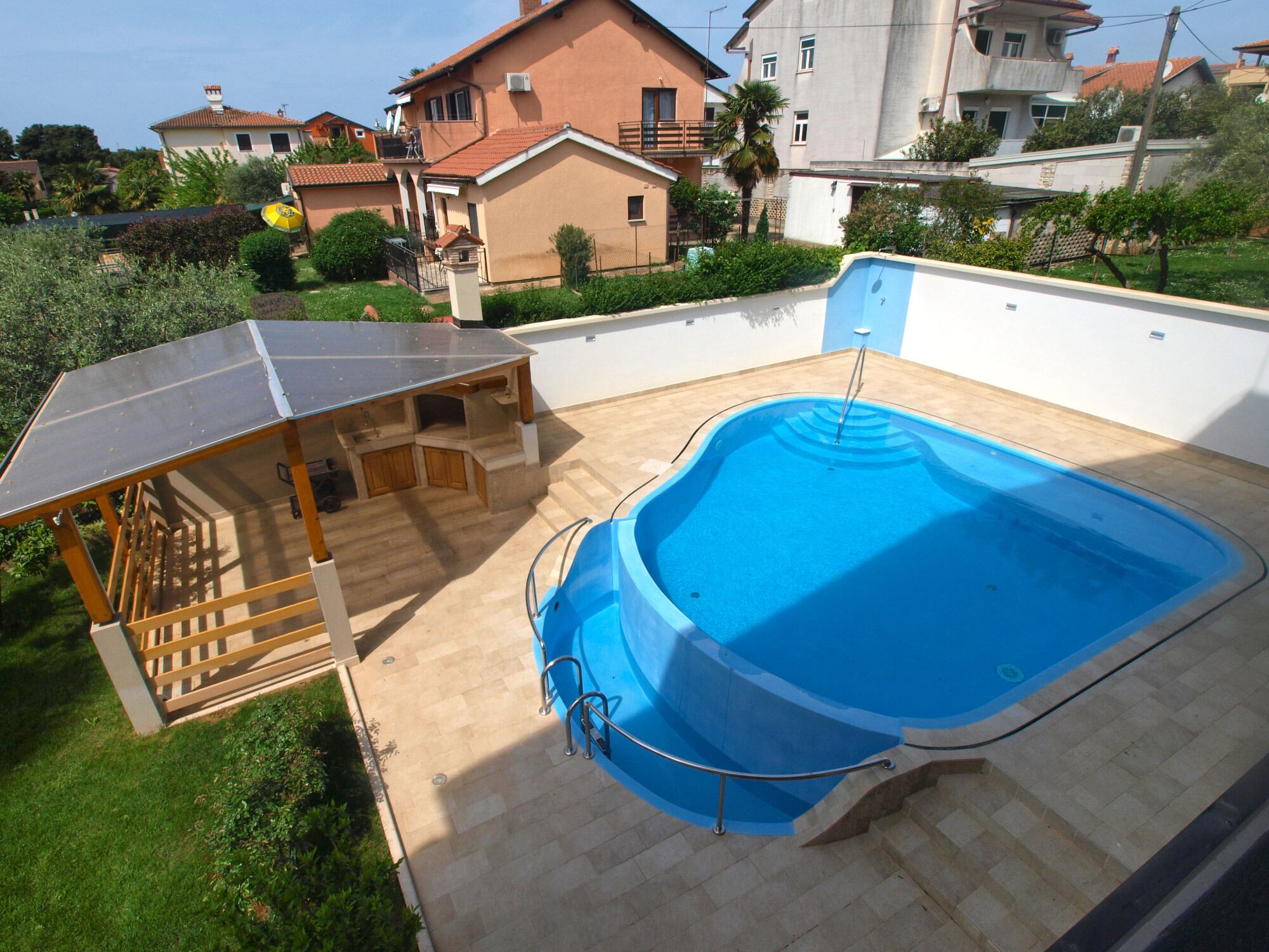 A rooftop swimming pool with a sunshade structure, a small outdoor kitchen, and a view of surrounding houses.