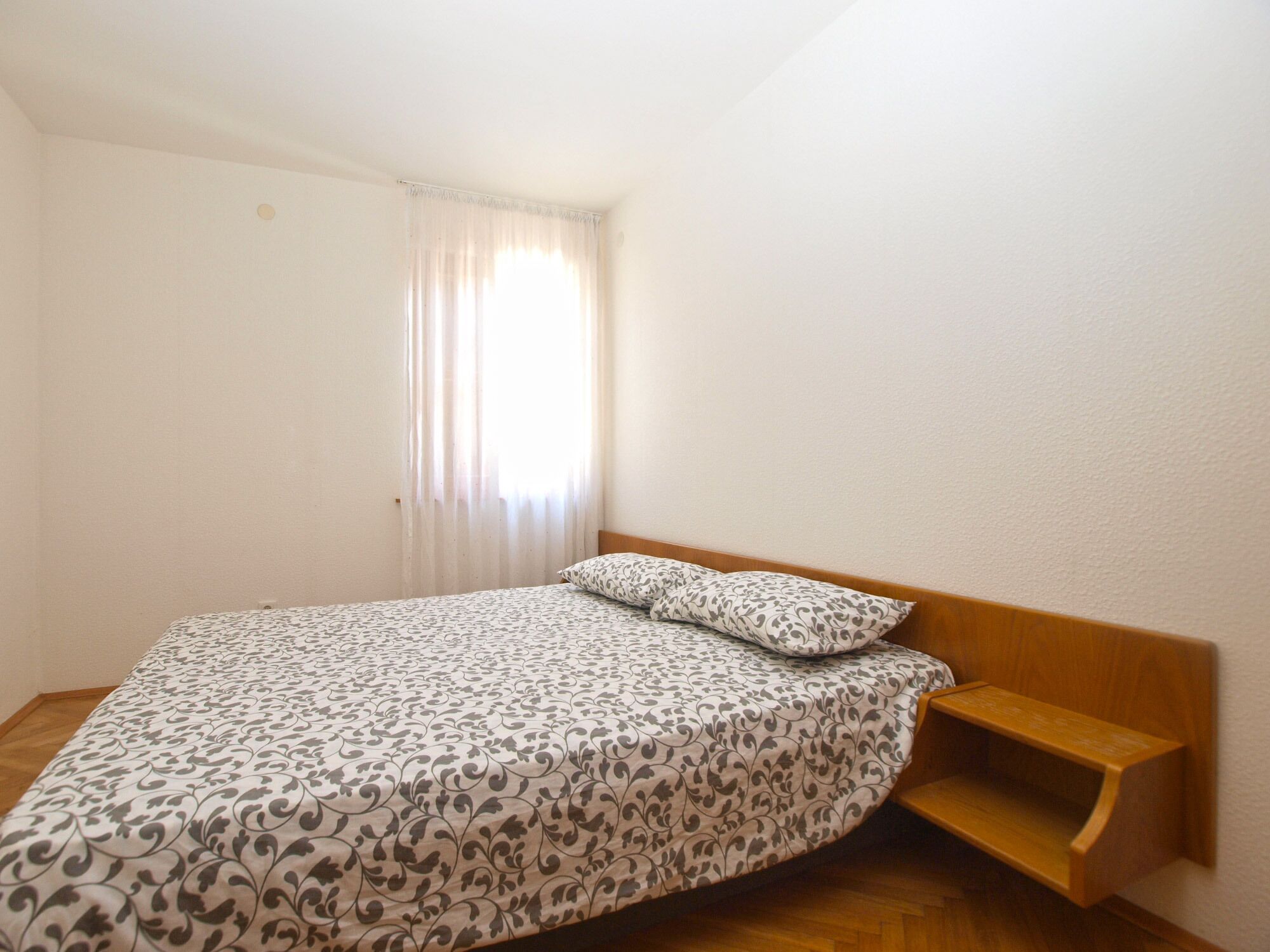 Uma cama de solteiro com colcha e almofadas estampadas, cabeceira de madeira e uma pequena prateleira de madeira.
