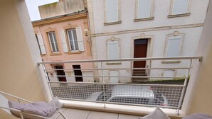 Een balkon met twee witte stoelen en een witte tafel, met uitzicht op een straat met gebouwen en een geparkeerde auto.