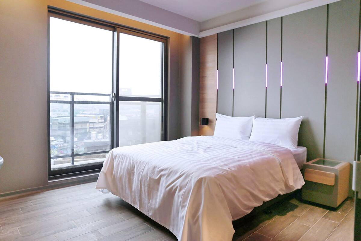 Une chambre d’hôtel moderne avec une grande fenêtre, un lit soigneusement fait, une table de chevet et un luminaire fixé au mur.