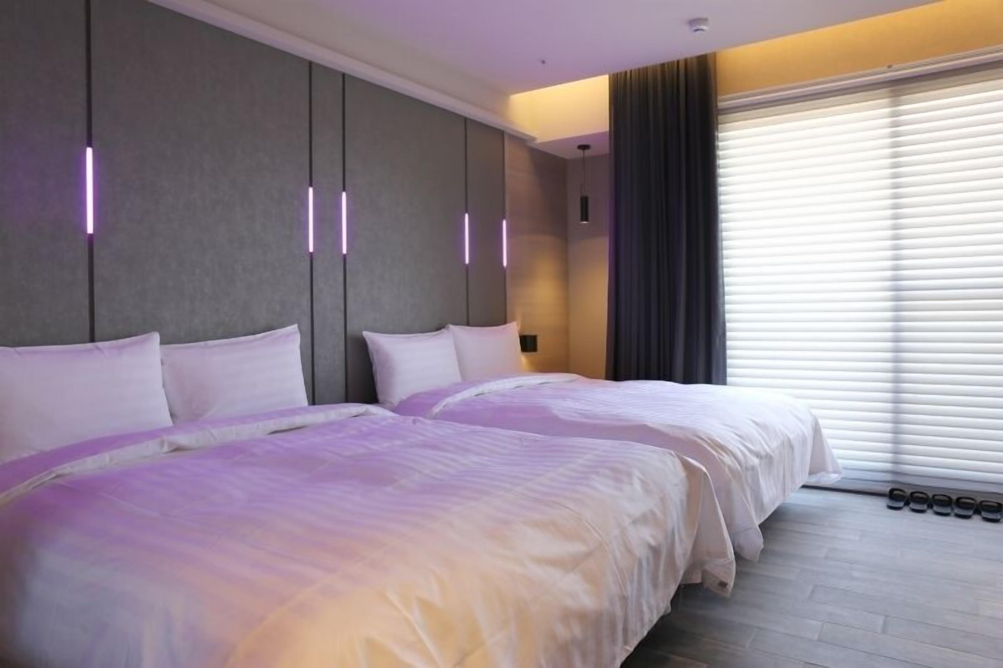 A hotel room with two beds, a large window with blinds, and a shoe rack by the window.