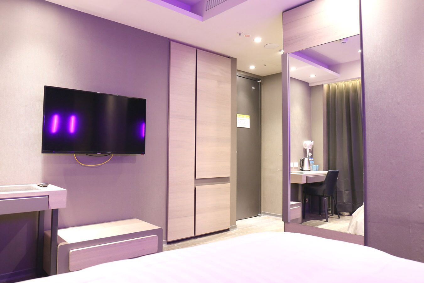 A modern hotel room with a flat-screen TV, a bedside table, a desk, and a mirror.