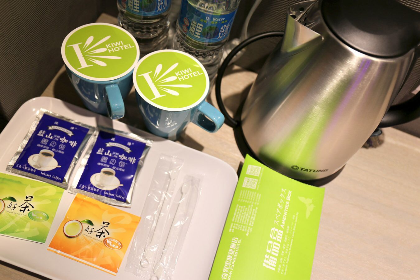 A tray with instant coffee packets, two blue mugs with a hotel logo, a stainless steel electric kettle, and a green amenity box.