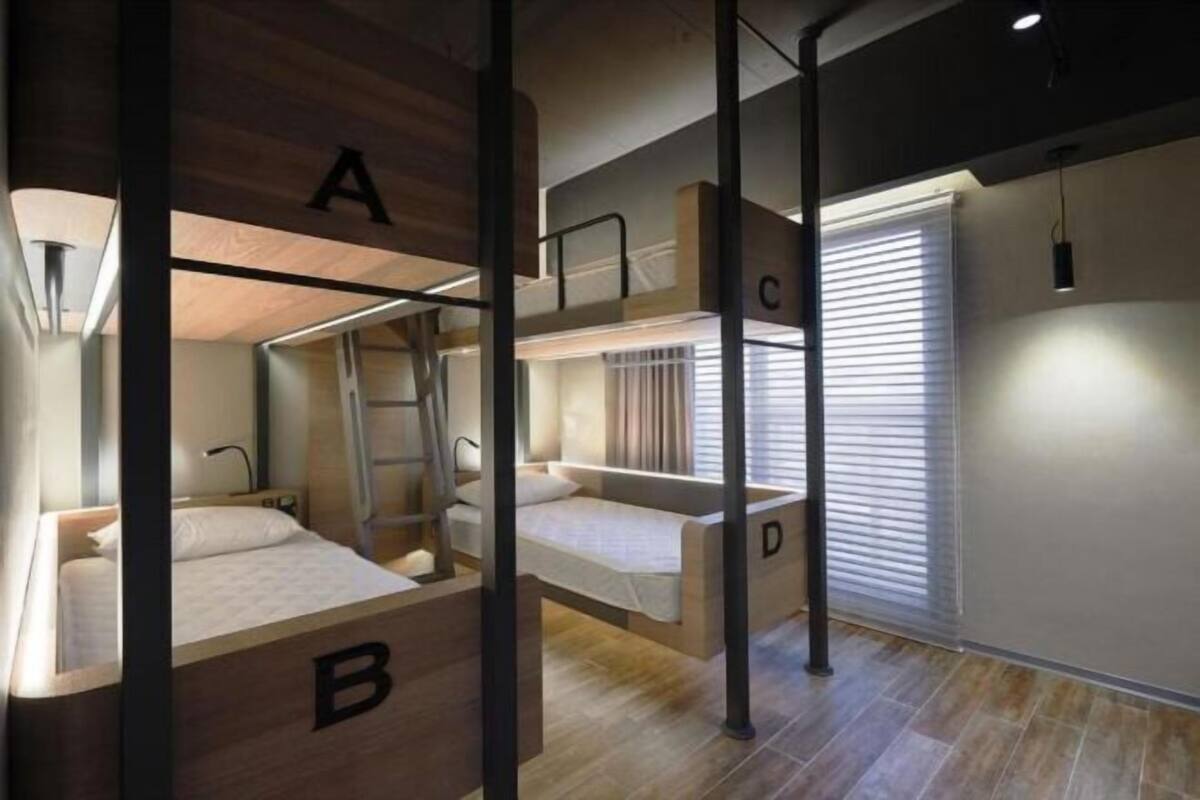 Une chambre d’hôtel moderne équipée de lits superposés étiquetés A, B, C et D, d’une échelle et d’une fenêtre avec des stores.