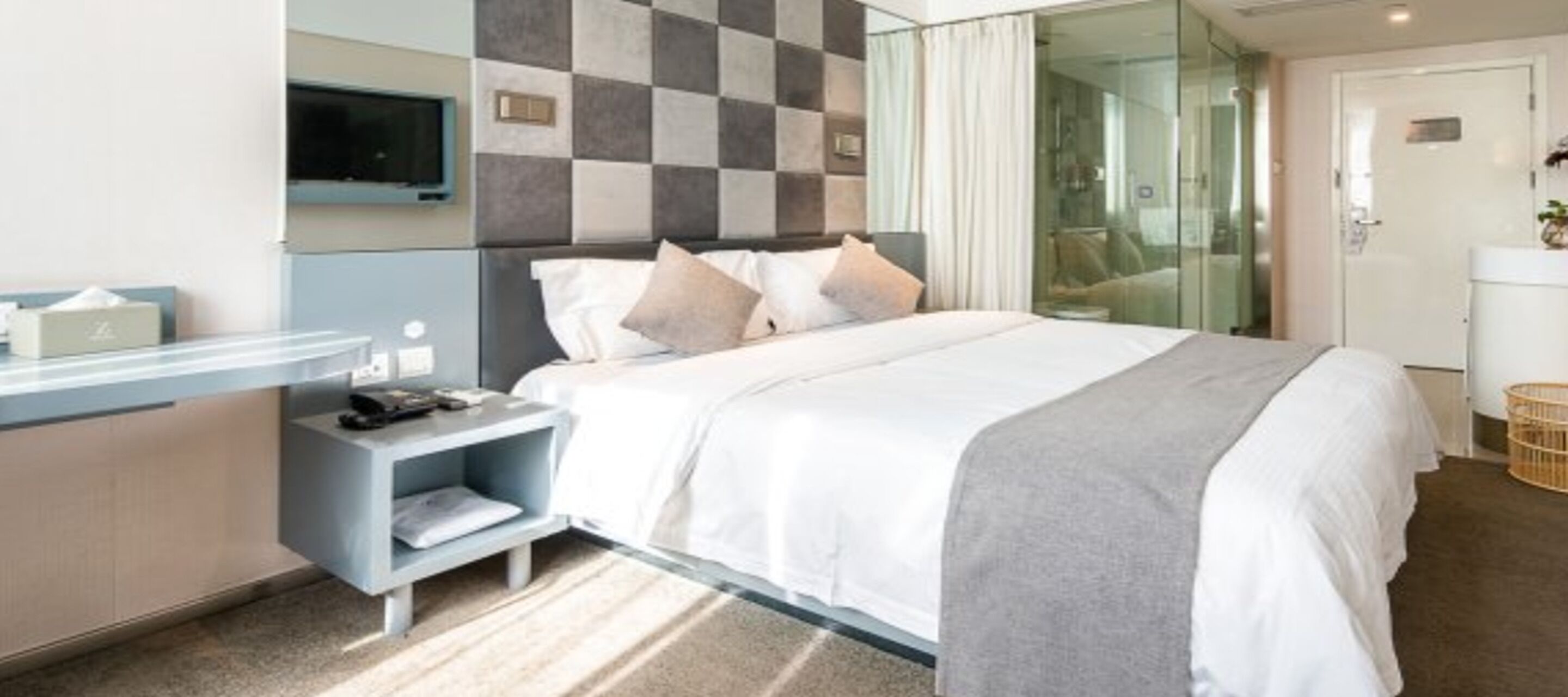 A modern hotel room with a large bed, a bedside table with a lamp, a flat-screen TV, and a glass partition wall.